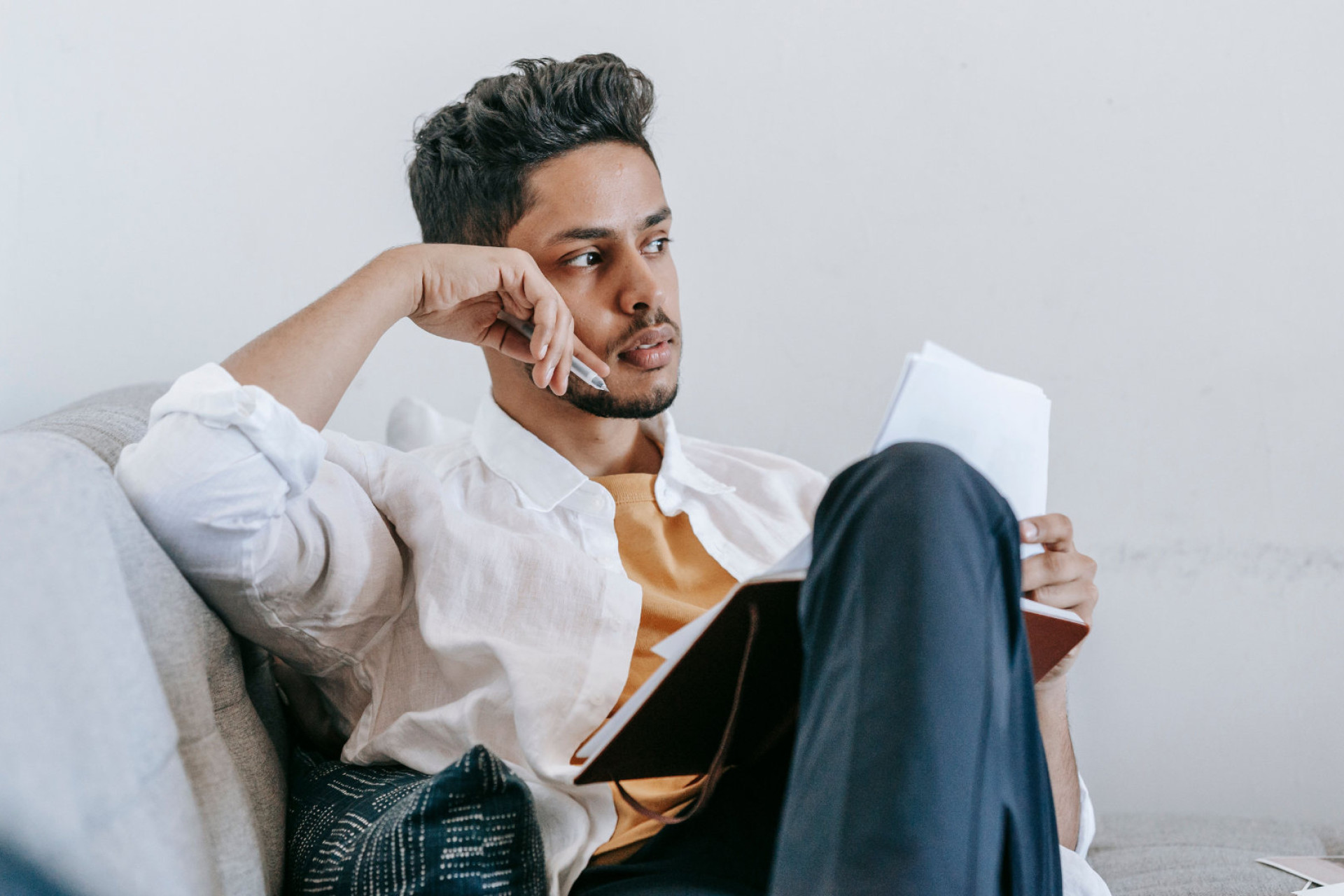 Man in wit overhemd zit op een bank met een notitieboek in zijn handen en kijkt peinzend opzij.