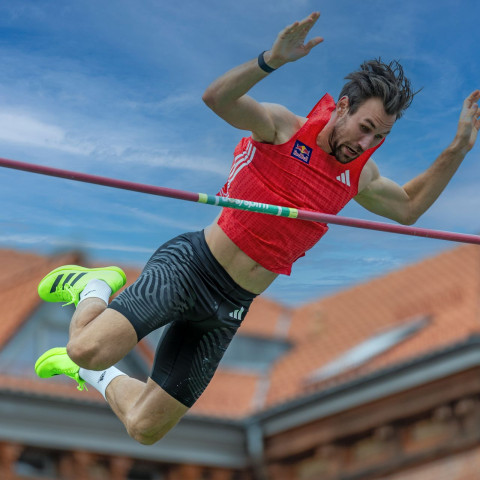 Een atleet gaat met gespannen gezicht net over de lat tijdens een polsstokhoogsprong.